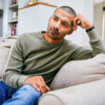 A young man sitting on his couch with his hand on his head, reflecting on the personal impact of common misconceptions about herpes.